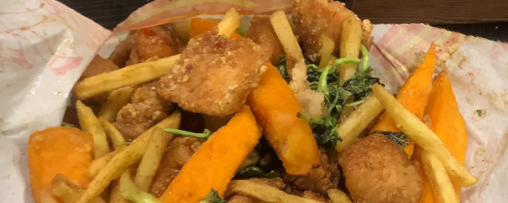 An overhead phone photo of a fried chicken order on paper — bite-sized fried chicken, sweet potato wedges, skin-on fries and fried broccoli, all in one pile