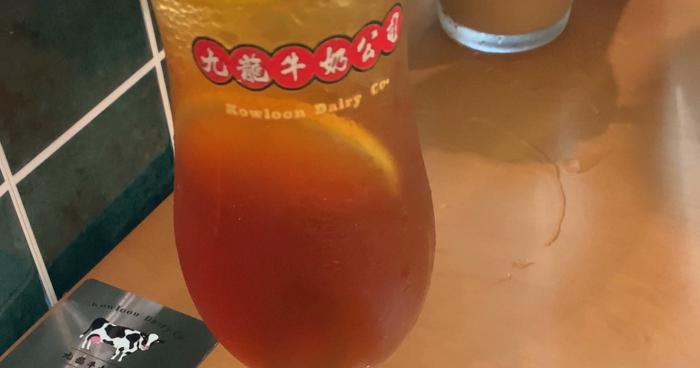 A tall glass of Hong Kong iced lemon tea with lemon slices on a wooden cha chaan teng table, green tiled wall behind it