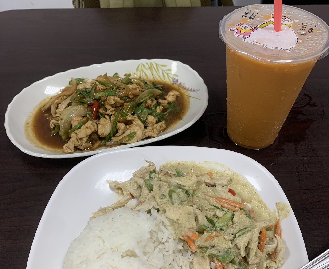 A Thai eatery table: a plate of stir-fried chicken with basil, green curry chicken over white rice, and a tall glass of Thai iced tea